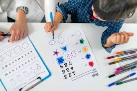 Bir çocuğun okul öncesi gelişimini ölçen bir test yaptığı an. Çocuk, şekilleri ve renkleri içeren bir kağıda renkli kalemlerle çalışıyor, yanında bir yetişkin rehberlik ediyor.